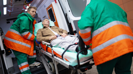 Paramedics in green and orange uniforms transport a male patient on a stretcher into an ambulance....