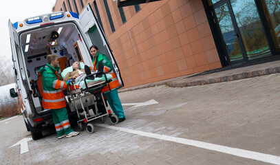 Naklejka premium Paramedics in green and orange uniforms transport a male patient on a stretcher towards a hospital entrance from an ambulance. The scene emphasizes emergency medical response and patient care.