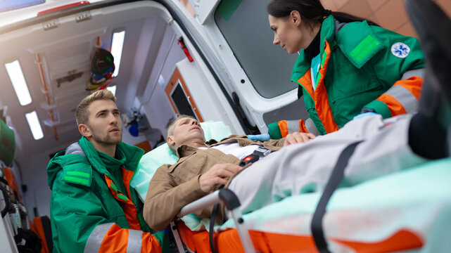 Paramedics in green and orange uniforms attend to a male patient on a stretcher, preparing him for transport inside an ambulance. The scene captures emergency medical care and teamwork in action.
