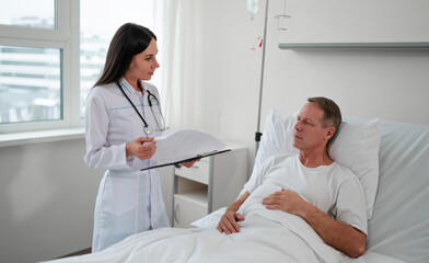 Obraz premium A female doctor reviews medical notes with a male patient resting in a hospital bed. The patient listens attentively while the doctor stands by with a clipboard. The room is bright and professional.