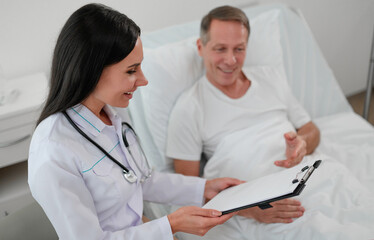 Fototapeta premium Smiling female doctor reviews notes on clipboard with a male patient lying in a hospital bed. The patient is engaged and relaxed. The bright, clean room emphasizes professional care and communication
