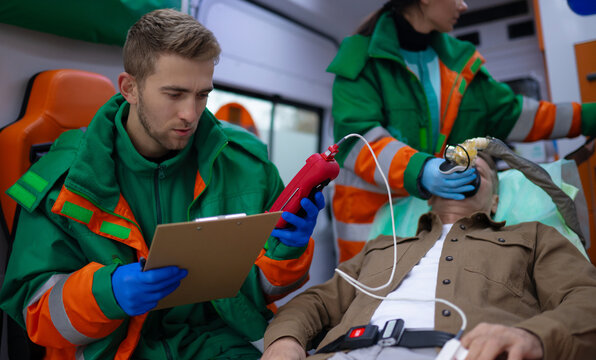 Paramedics in green and orange uniforms tend to male patient in an ambulance. One checks vitals with clipboard while the other administers oxygen. The scene shows emergency medical care in progress