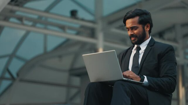 Busy businessman in black jacket sitting alone with laptop on laps and working in uncomfortable position. Indian businessman concentrating on result and answering question from distant colleague.