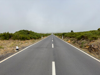 An empty, straight, paved road with a white dividing strip, stretching to a misty horizon. Madeira, Portugal.
