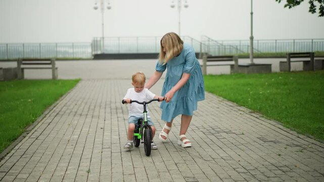 A mother is gently guiding her young son as he rides his bicycle along an interlocked pathway, the boy appears focused, while his mother helps to steady him