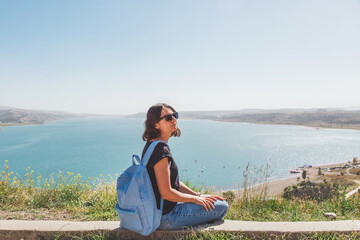 one white brunette young girl in sunglasses tourist sitting on a hill in front of a large reservoir lake with a backpack at Tbilisi Sea in Georgia