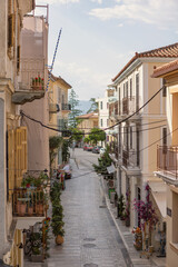 Wander through a picturesque Nafplio street adorned with vibrant blooms and inviting balconies, leading toward a serene coastal view illuminated by the midday sun.