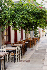 Colorful tables and chairs are arranged along a tranquil street in Nafplio, creating an inviting atmosphere for relaxed dining amidst vibrant foliage.