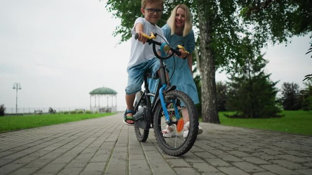 A woman wearing a blue gown helps her son maintain balance as he rides his bicycle down a brick path in a park