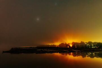 A calm, misty riverbank at night, with boats resting along the shore. Warm orange and yellow lights illuminate the distant trees and reflect on the water, creating a tranquil, atmospheric scene.