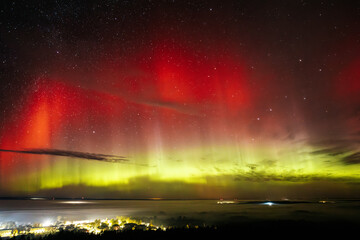 A vivid aurora borealis with striking red and green hues lights up the starry night sky above a town. The vibrant celestial display contrasts beautifully with the illuminated town below, Zilaiskalns.