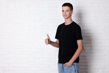 Teenage boy wearing black t-shirt and showing thumbs up near white brick wall, space for text