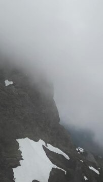 Vertical video of the side of a fog and cloud covered mountain top, with patches of snow on the ground. British Columbia, Canada.