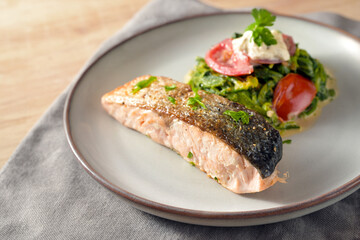Salmon fillet roasted with a crispy skin, vegetables from cream spinach, onions, tomatoes and parsley garnish, healthy fish meal on a gray plate, low carb diet, copy space