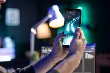 Closeup of a person holding a smartphone in front of a laptop, engaged in an online video game. Detailed view of mobile device displaying entertainment graphics, played by an individual.
