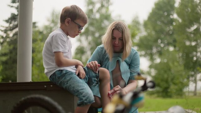 A mother squats as she carefully cleans her son's bleeding leg, the child looks upset while sitting on a bench