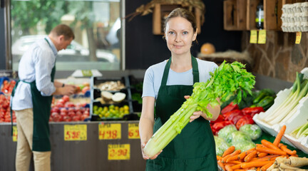 Woman seller of fruit and vegetable store works near showcase with garden-stuff, sorting greenery celery, checking goods.