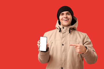 Young man in stylish puffer jacket and hat with mobile phone on red background