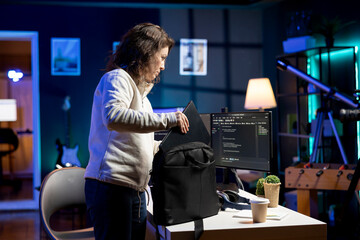 System administrator packing backpack, leaving office after implementing security measures, protecting company systems and data. IT staff member ending work shift after finishing tasks