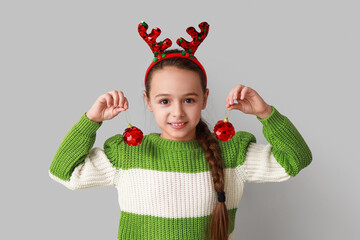 Cute little girl in reindeer horns with Christmas balls on grey background