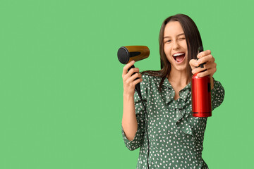 Happy young woman with hair dryer and spray on green background