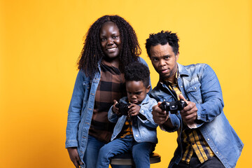Cheerful african american dad playing video games with his son on console, using controller to have fun against yellow background. Happy positive young family enjoying their leisure time.