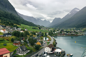Alusund, Norway 15 Aug 2024 - A scenic fjord surrounded by towering mountains, with a charming village along its shore. Lush green fields and winding roads complete the idyllic landscape