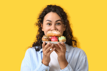 African-American woman with tasty eclairs on yellow background