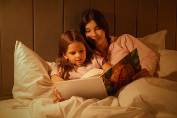 Happy mother and her little daughter reading fairy-tale in bed at night