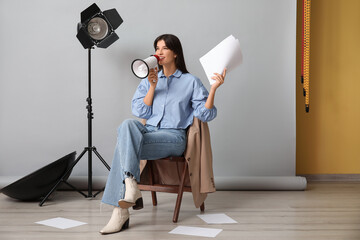 Young happy female film director with megaphone, script and studio lighting sitting on chair...