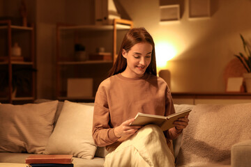Beautiful young happy woman reading book on sofa in evening at home