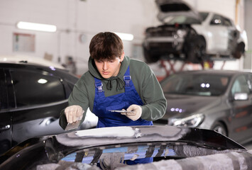 Skilled young auto body technician in garage, wearing blue overalls and green hoodie, applying filler to damaged car bumper, preparing for painting..