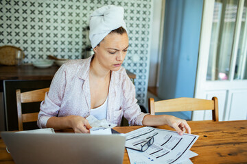 Serious woman working from home with laptop and bills in kitchen