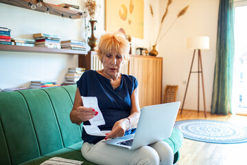 Elderly woman reviewing receipts and using a laptop while sitting on a couch