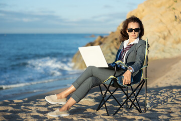 Fototapeta premium Relaxed woman in business attire with white shirt, loose tie and sunglasses working on laptop while sitting on quiet ocean beach on sunny day, blending work with peaceful escape in nature