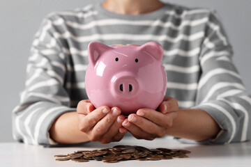 Woman holding piggy bank over white wooden table with coins, closeup