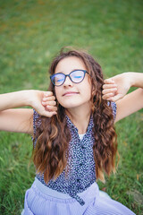 Portrait of girl in glasses with curly hair sitting in park on grass. Concept of learning in nature