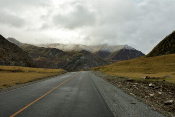 A straight asphalt road crosses a picturesque valley towards high mountains on a cloudy and overcast autumn day.