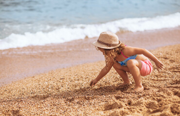 Happy kid girl sitting in summer clothing, straw hat and playing with sand of the beach near blue sea background. Happy