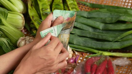 Close-up of hands counting Indonesian Rupiah while purchasing fresh vegetables from a market. The scene captures vibrant greens and local produce, symbolizing daily life and commerce. - Powered by Adobe