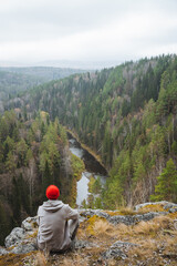 A man is sitting on a large rock while he overlooks a peaceful, serene forested valley that features a gently flowing river below