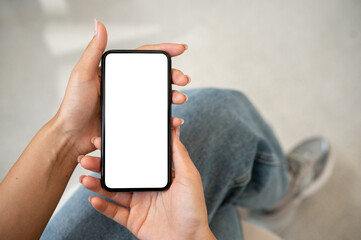 A woman in jeans sitting indoors and using her smartphone, with a white screen mockup displayed.