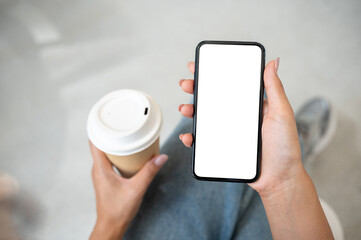 Top view of a woman in jeans sitting in a cafe, holding a coffee cup while using her smartphone.