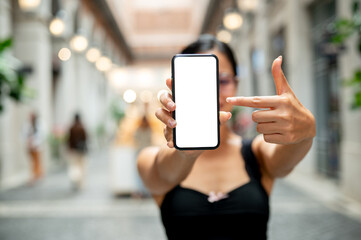 A woman pointing at a smartphone, showing it to the camera while standing in a market or mall.