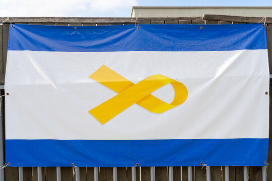 Close-up of a blue and white flag featuring a prominent yellow ribbon symbol, displayed on a fence. The flag represents support and awareness for the hostages in Israel.