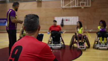 Rear view of a male wheelchair rugby player observing teammates during a team strategy session on the court.