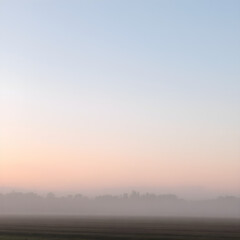 Fototapeta premium A soft, misty morning sky with pastel tones of pink and blue, with the fog rising from the fields and trees barely visible in the distance 