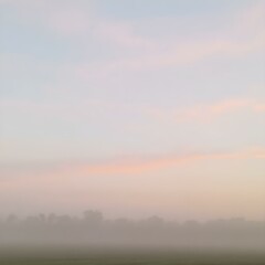 Fototapeta premium A soft, misty morning sky with pastel tones of pink and blue, with the fog rising from the fields and trees barely visible in the distance 