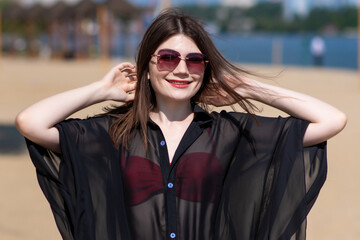 Beautiful, attractive, happy girl posing on a sandy beach on a sunny day in a red swimsuit, sunglasses and a black beach tunic. Beachwear