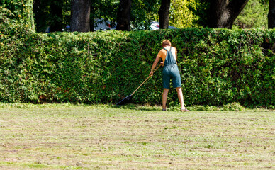 A woman in a green apron is walking through a hedge with a rake in her hand